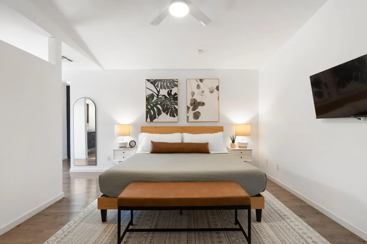 A cozy bedroom setup with a bed topped with plush, decorative pillows. Framed wall art, nightstands with lamps, and a mirror enhance the sense of space and luminosity. The room is bathed in natural light, suggesting a bright, uplifting start to the day.