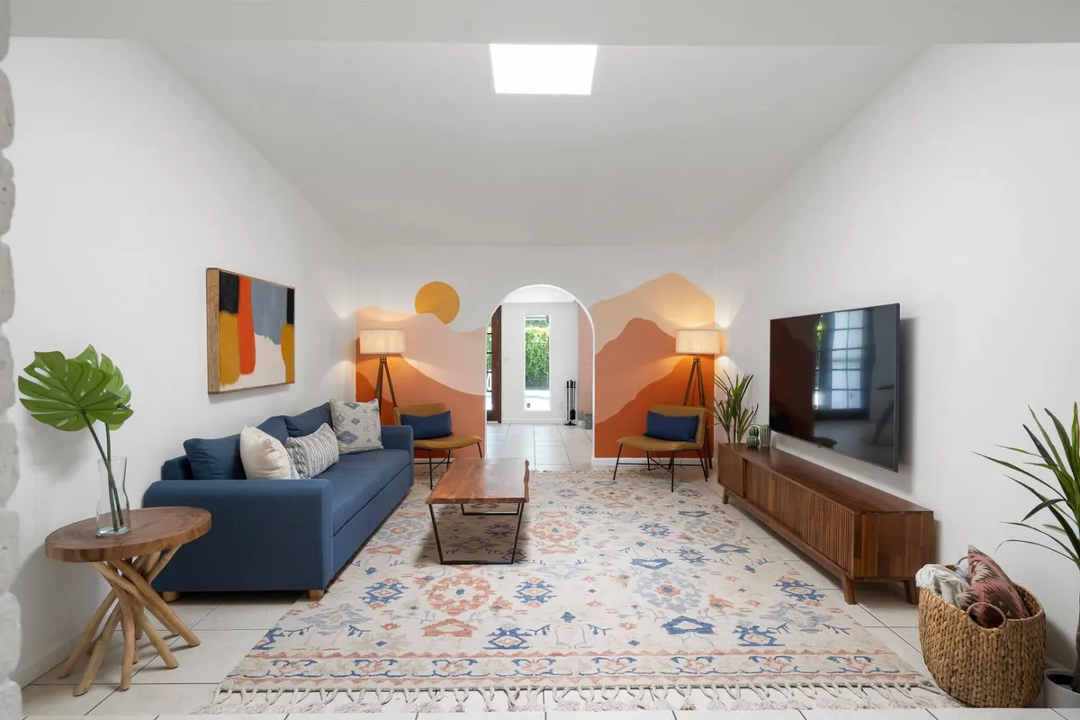 This image showcases a welcoming living room with a comfortable sofa, eye-catching mountain mural, television, coffee table, and abundant natural light streaming in, emphasizing a space designed for relaxation, conversation, and group activities.