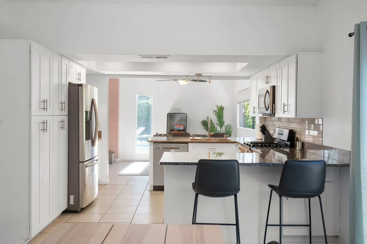 A vibrant kitchen scene highlighting a central island with seating, stainless steel appliances, large windows letting in natural light, and a green plant adding a fresh touch. The open design promotes interaction and easy entertaining.