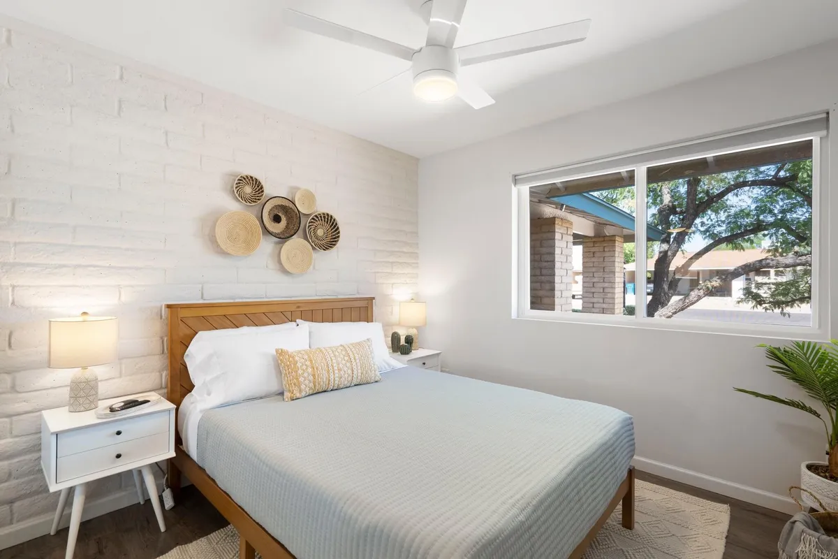 A tranquil bedroom featuring a comfortable bed dressed in soft, inviting bedding. Decorative wall art, nightstands with lamps, and natural light streaming through the windows create a peaceful retreat. The thoughtfully arranged space feels personal and cozy, ideal for resting or unwinding with a book.