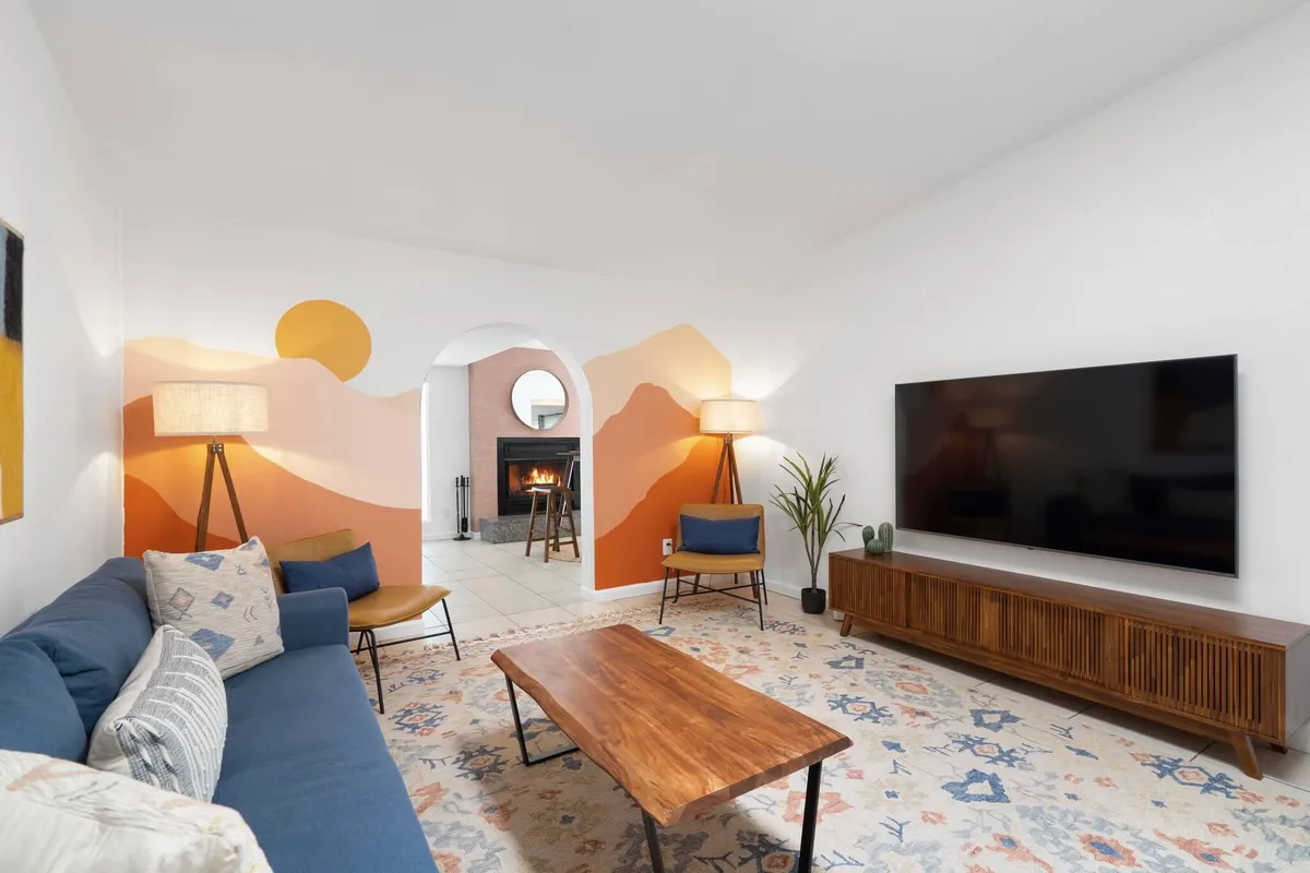 The image showcases a warm, inviting living room with a vibrant, colorful wall mural as the centerpiece. A sleek wooden coffee table sits in front of a modern television, flanked by decorative lamps that cast a cozy glow. The arrangement suggests a space perfect for relaxing, socializing, or enjoying quiet moments, with sunlight streaming in to brighten the room.