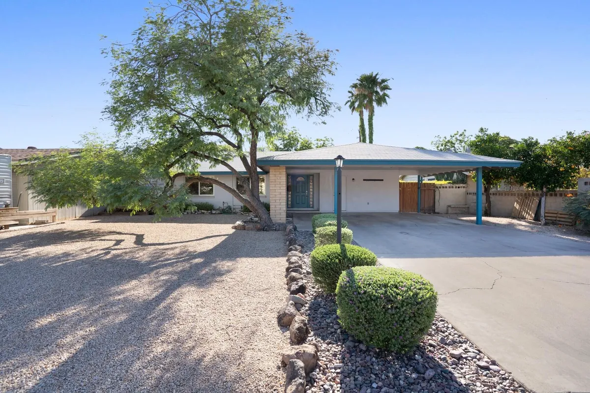 The scene features a view of the property's exterior with a tree, carport, landscaped shrubs, and a gravel driveway. The inviting, homey environment hints at a welcoming base for daily adventures and the tranquil neighborhood setting.